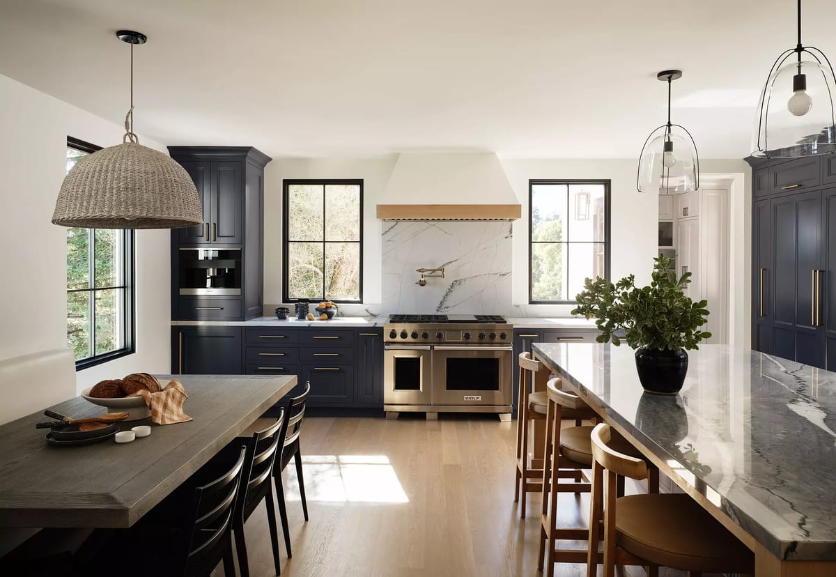 Kitchen with navy cabinets, marble island, six-burner stove, wooden dining table, and large windows for natural light