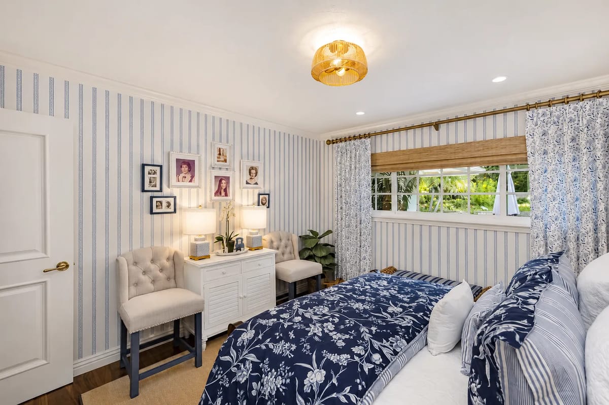 Bedroom with blue floral comforter, nightstands, cushioned chairs, striped wallpaper, and large window.