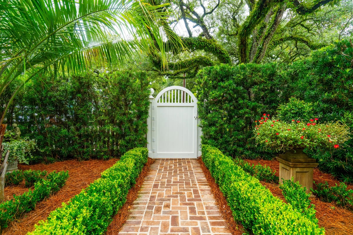 Garden pathway with hedges, flower pots, brick path, and a white wooden gate.