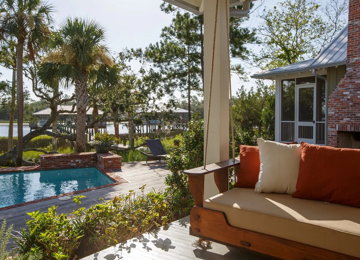 Patio with wooden bench, swimming pool, palm trees, and landscaped greenery overlooking water and dock.