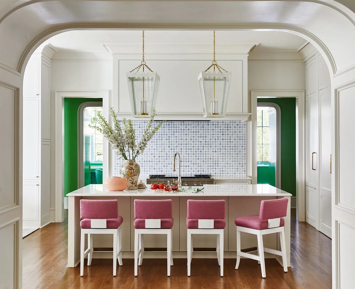 Kitchen with pink stools, white countertop, mosaic backsplash, green walls, and large windows providing natural light.