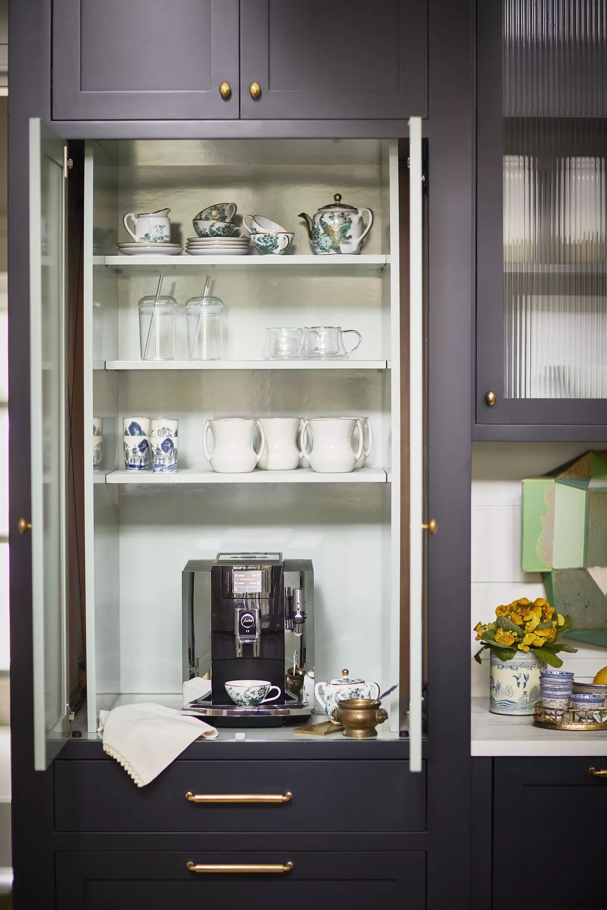 Kitchen display cabinet with tea sets, glassware, and coffee machine above a counter with flowers.