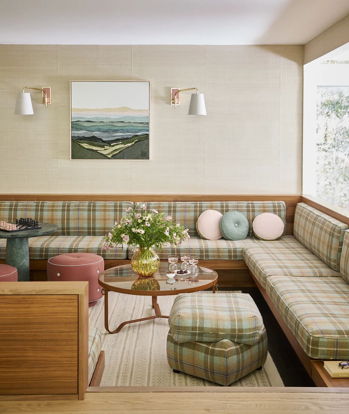 Living room with plaid sectional sofa, coffee table, cushions, flower vase, pink stools, wall sconces, and artwork.