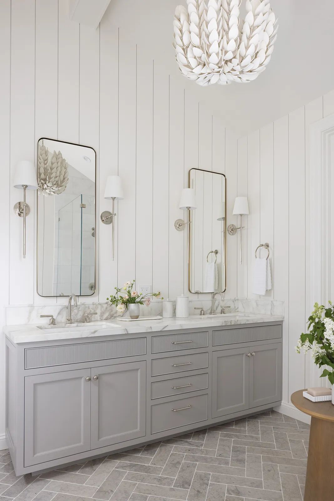Bathroom with gray vanity, marble countertop, mirrors, wall sconces, and herringbone-patterned flooring.