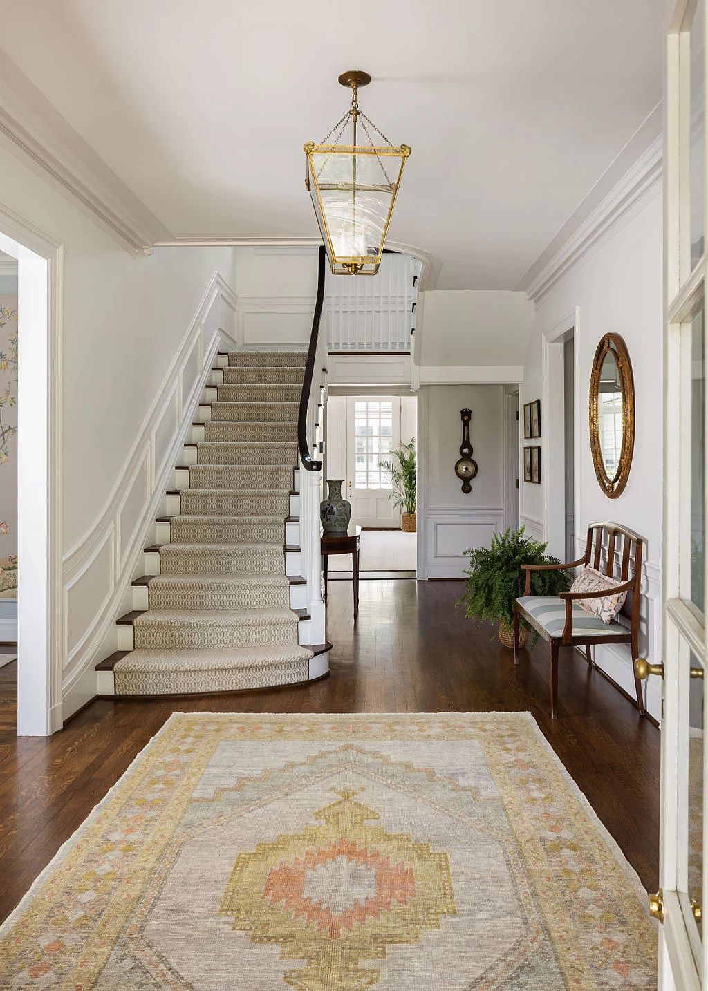 Foyer with staircase, area rug, bench, mirror, clock, and potted plants.