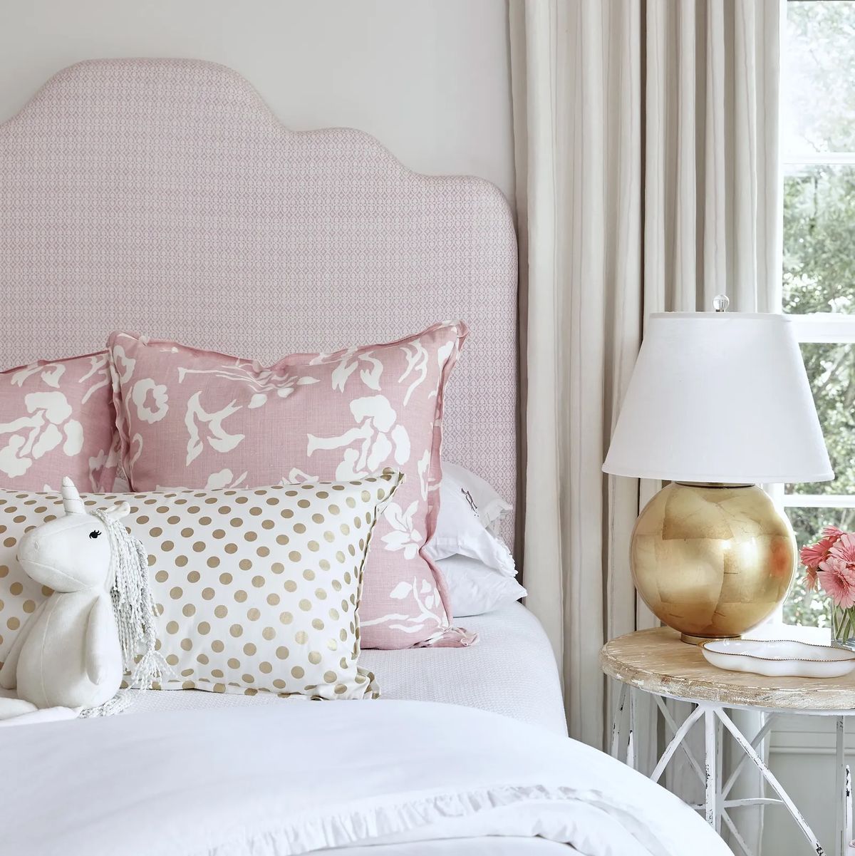 Bedroom with pink headboard, decorative pillows, white bedspread, side table, gold lamp, and vase of flowers.