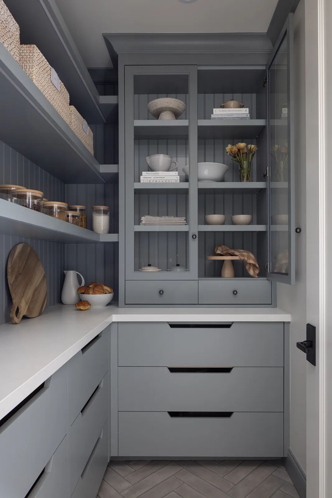 Butler's pantry with gray cabinetry, shelving, white countertop, glass jars, and herringbone flooring.