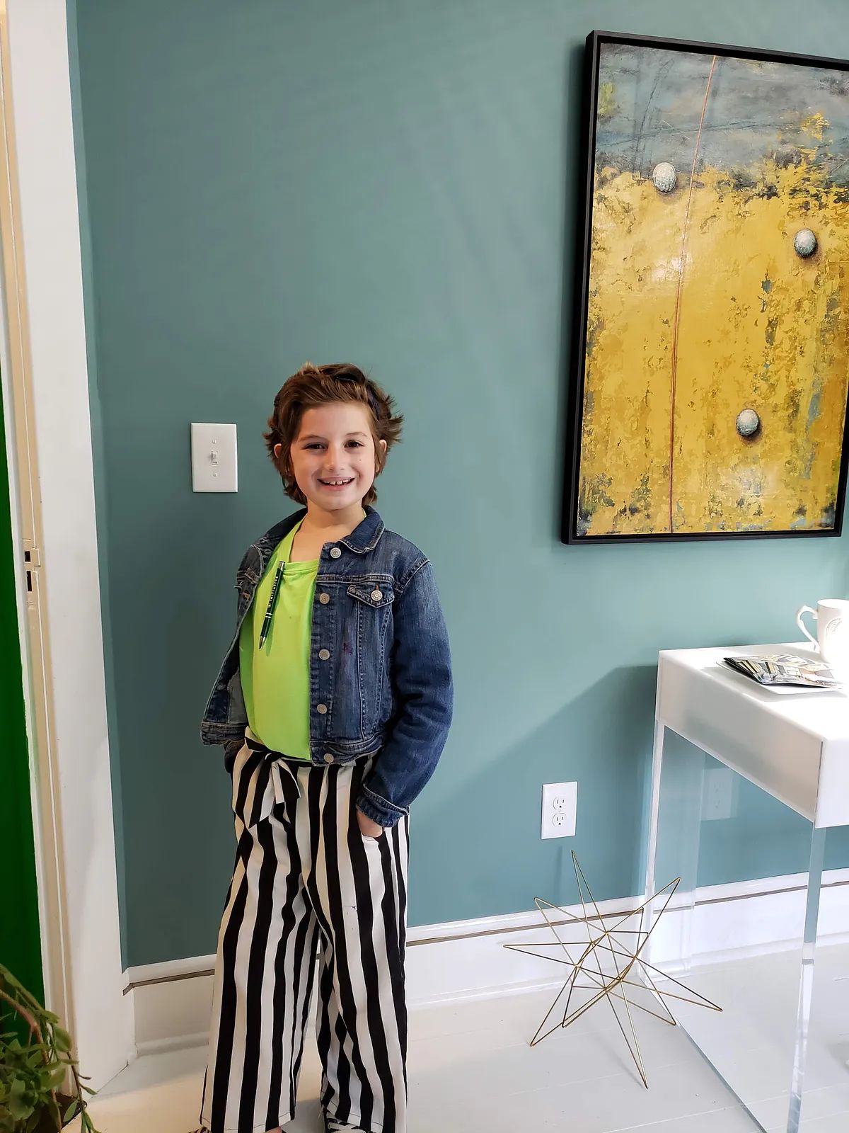 Small room with green wall, artwork, clear table, and a child in denim jacket and striped pants.