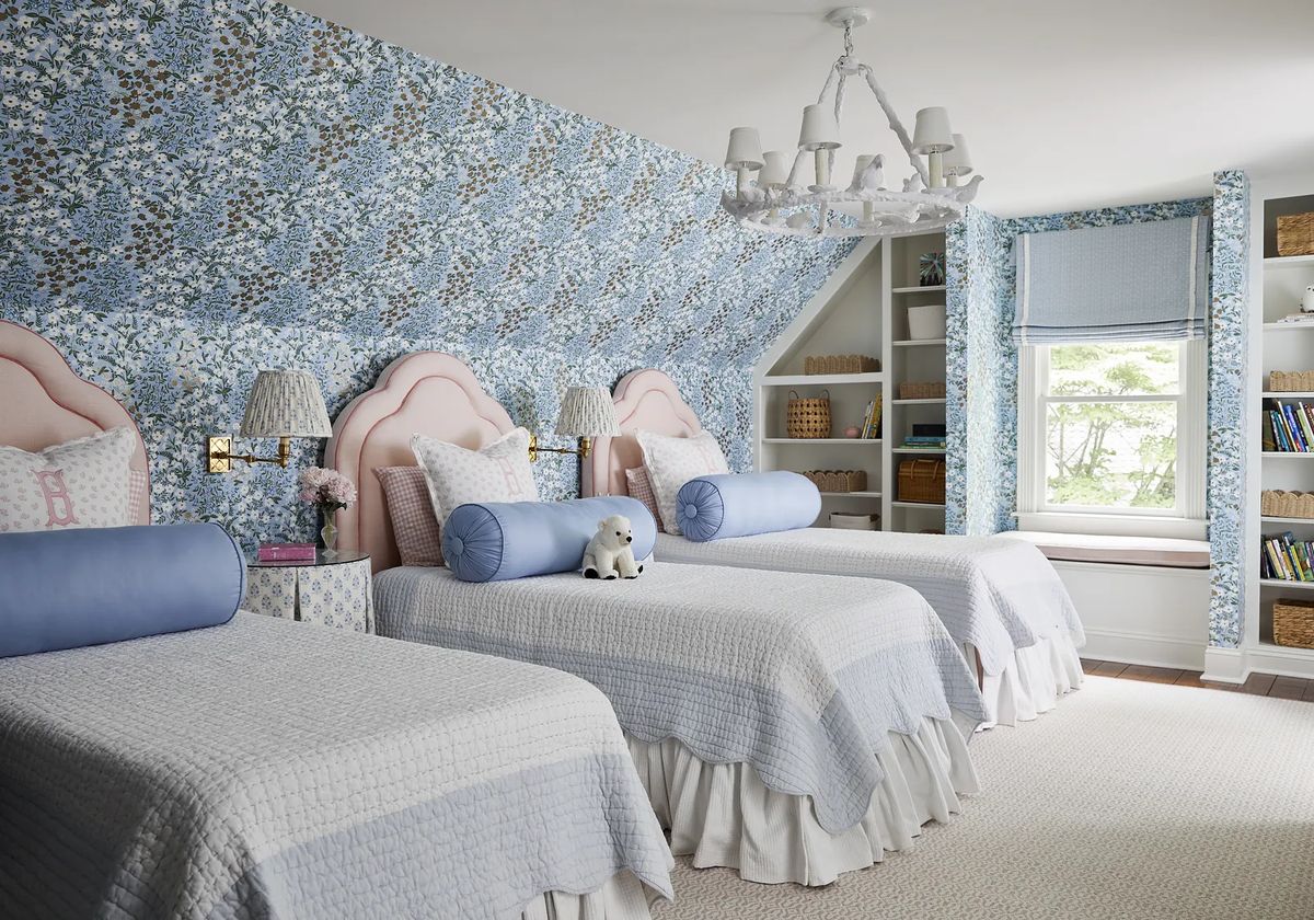 Bedroom with three twin beds, pink headboards, blue bolster pillows, patterned wallpaper, and built-in shelves