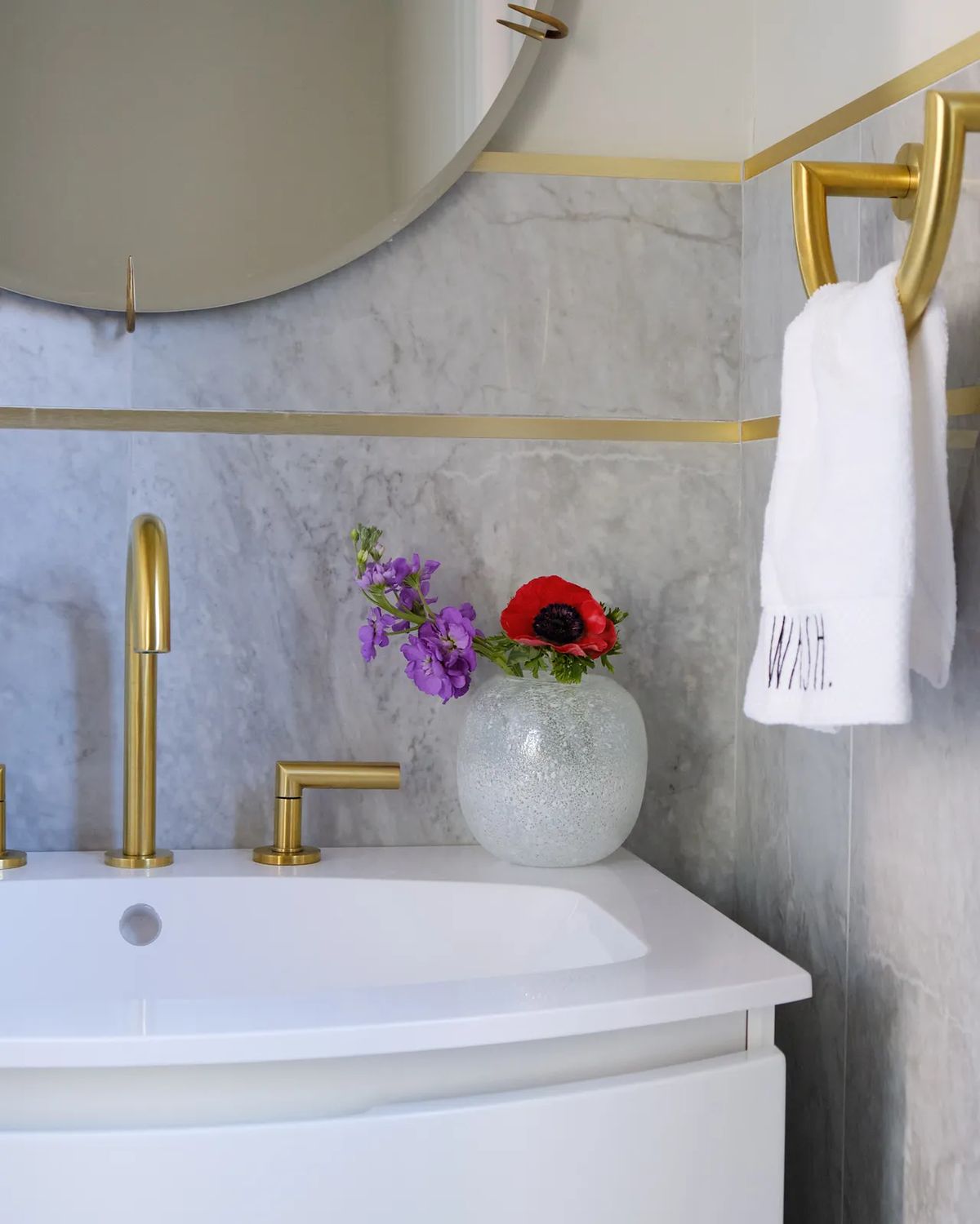 Bathroom with white sink, gold faucets, round mirror, marble wall, flowers, and towel holder.
