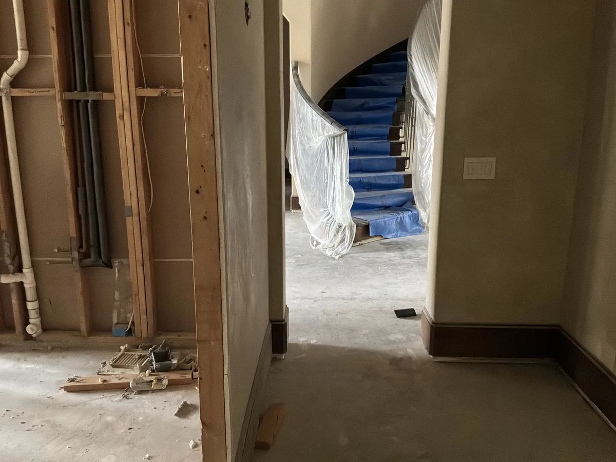 Hallway under construction with exposed plumbing, bare floor, and blue-covered staircase.