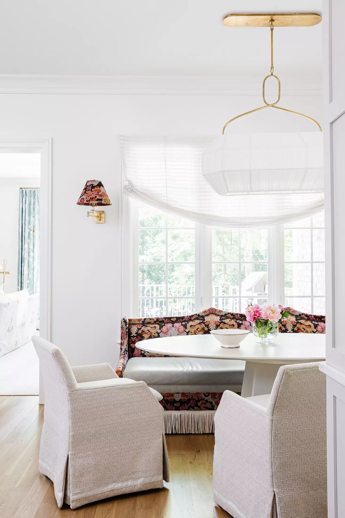 Dining room with round table, upholstered chairs, floral bench, and natural light from large windows.