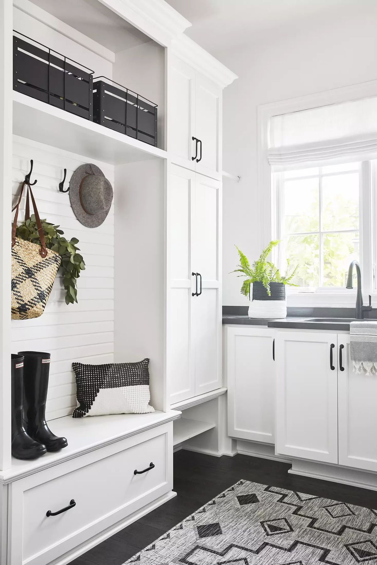 Mudroom with built-in cabinetry, sink, window, black boots, hat, and patterned rug.