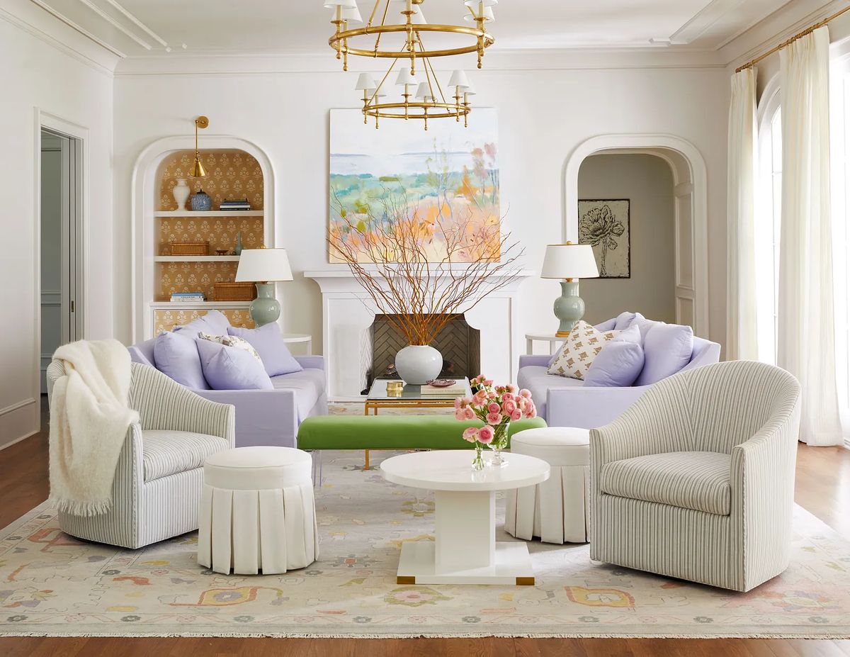 Living room with purple armchairs, green vases, white coffee table, ottoman, floral rug, painting, and large windows