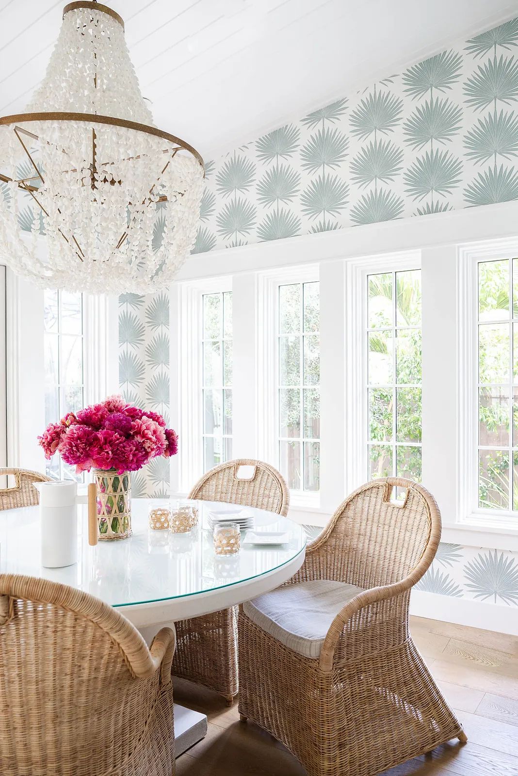Dining room with round glass table, woven chairs, pink flowers, and palm leaf patterned wallpaper.