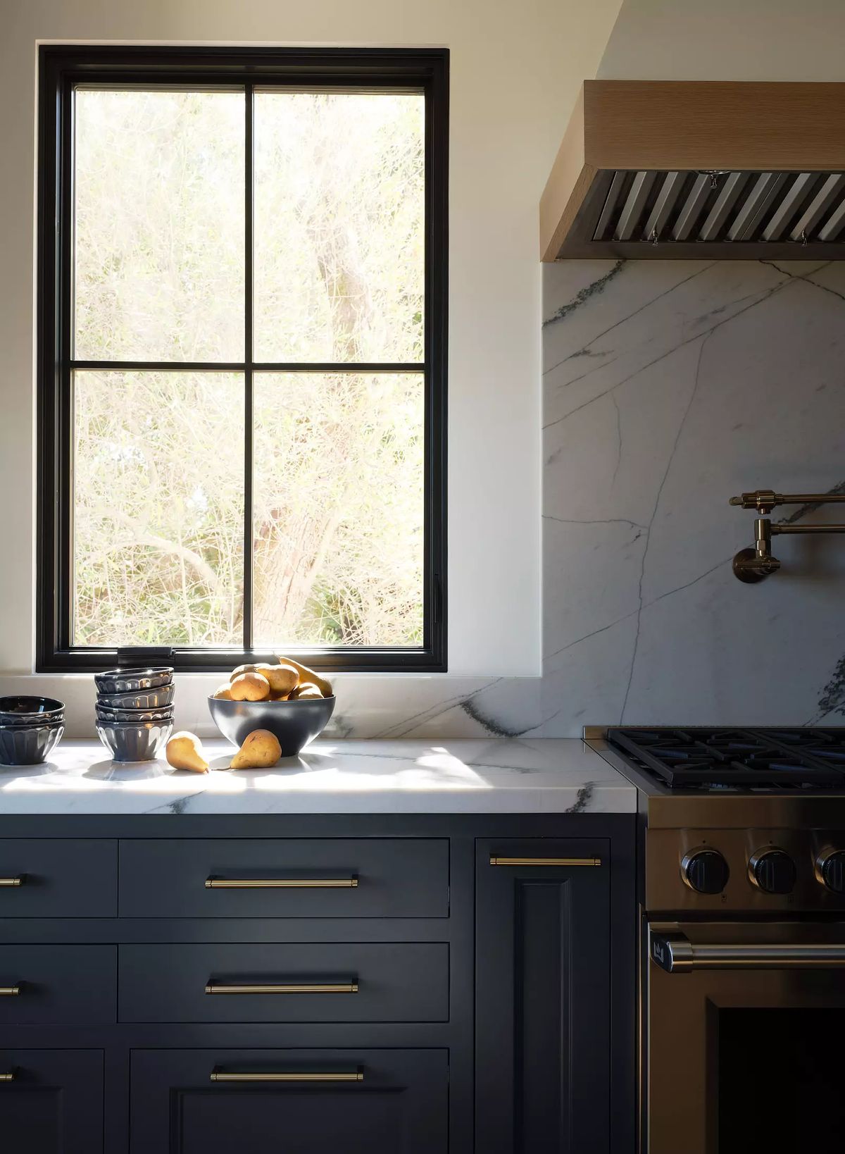 Kitchen with dark blue cabinetry, marble countertop, gas range, and natural light through a large window.