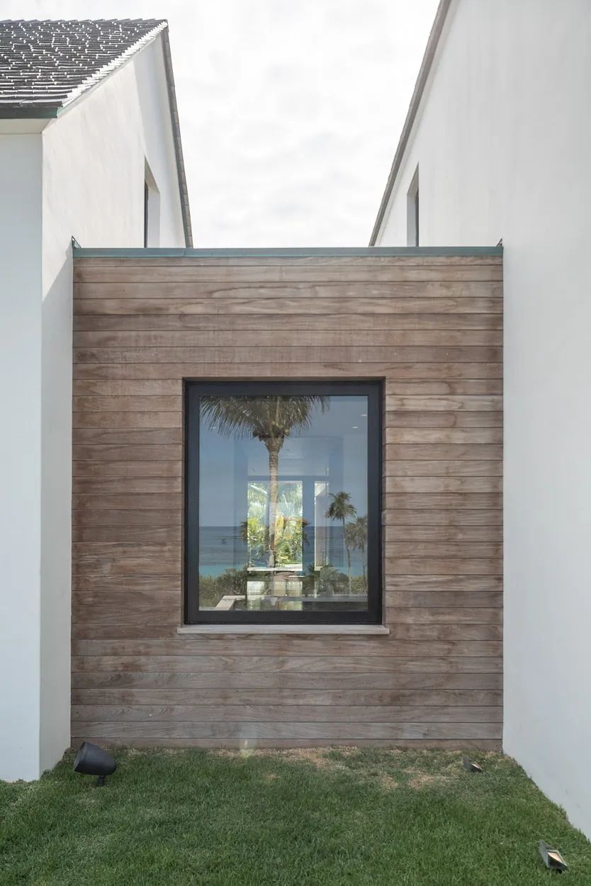 Exterior view with wooden wall, large window showing palm tree and seating, surrounded by white walls and grass