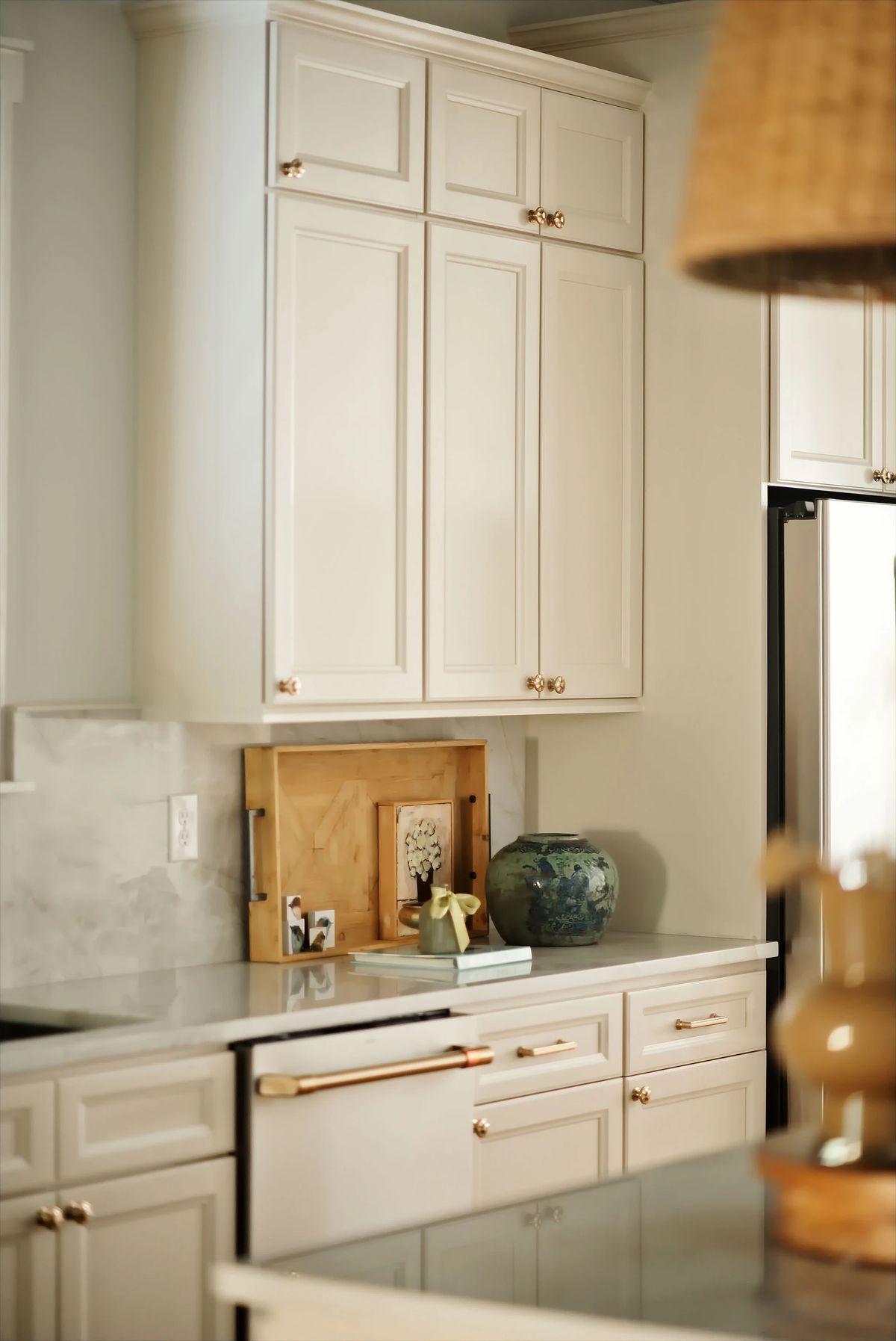 Kitchen with upper cabinets, gray countertop, stainless steel refrigerator, decorative items, and gold hardware.