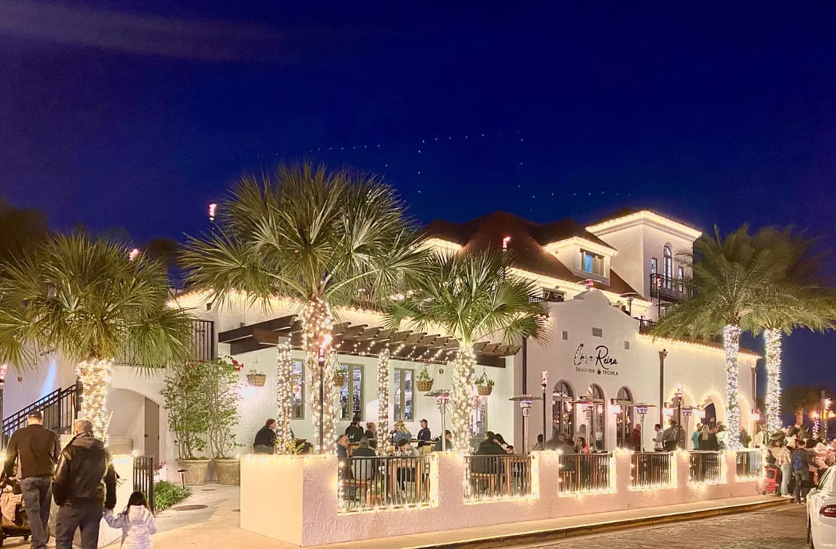 Exterior view of a building with white stucco, palm trees, string lights, and outdoor seating.