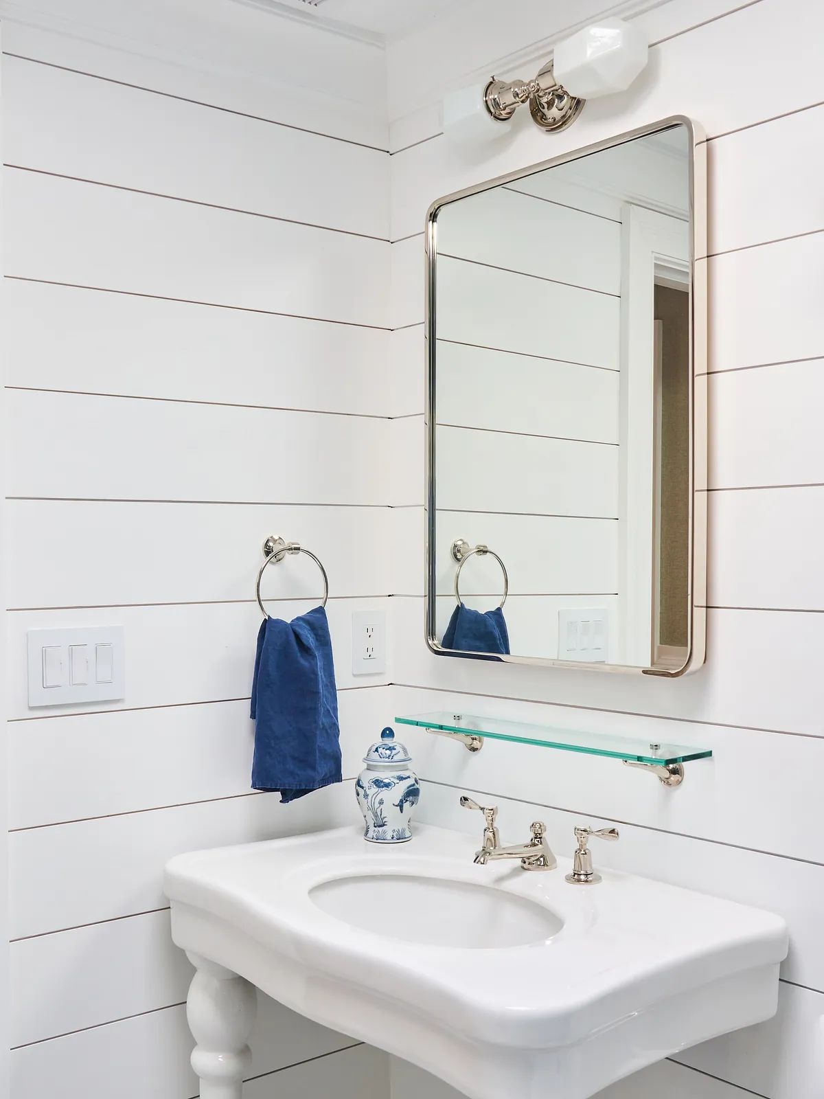 Bathroom with white pedestal sink, glass shelf, mirror, blue towel, and shiplap walls.