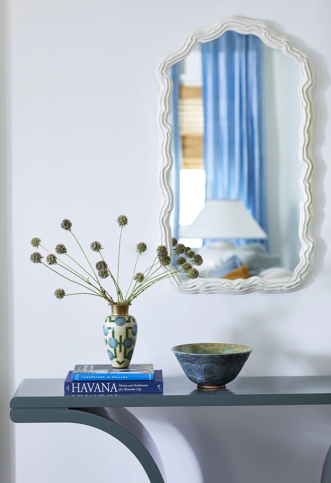 Console table with vase, books, ceramic bowl, and round mirror with blue curtains in the background.