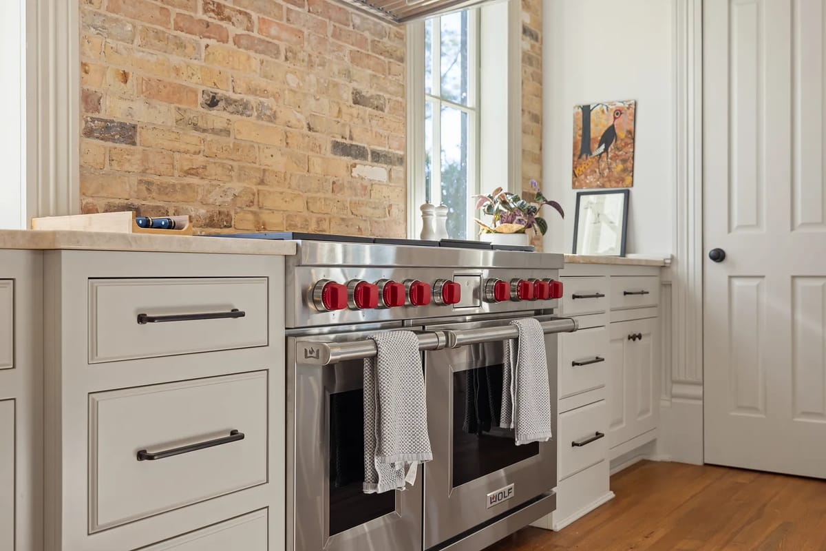 Kitchen with stainless steel range, white cabinetry, stone countertop, and exposed brick walls with large windows.