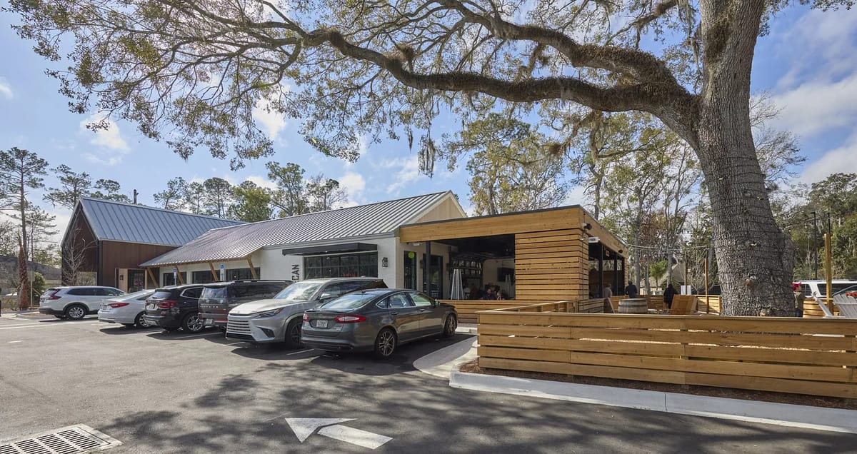 Outdoor commercial space with parked cars, wooden seating areas, and a large tree.