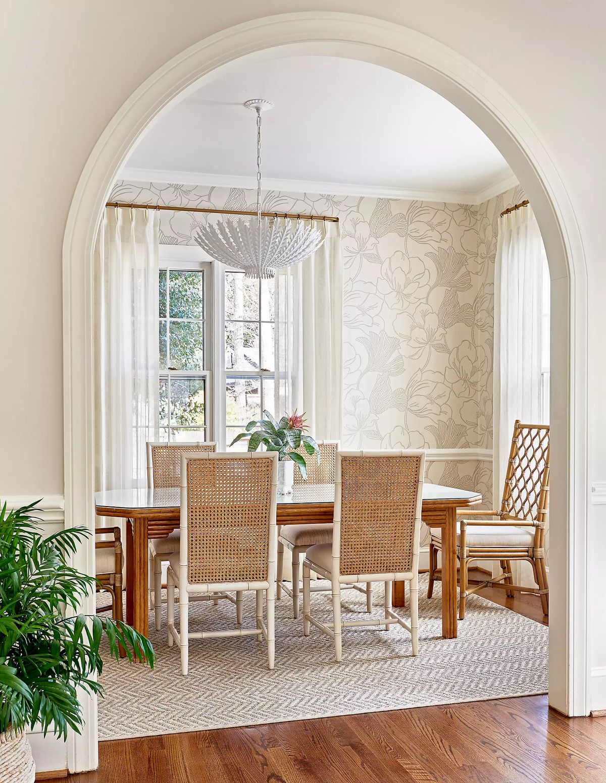 Dining room with wooden table, six woven chairs, floral wallpaper, large window, and pendant light