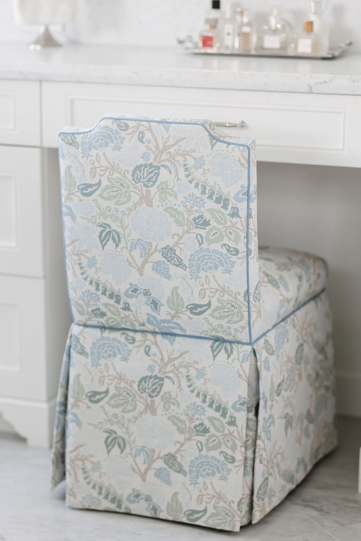 Chair with floral slipcover near white countertop with perfume bottles.