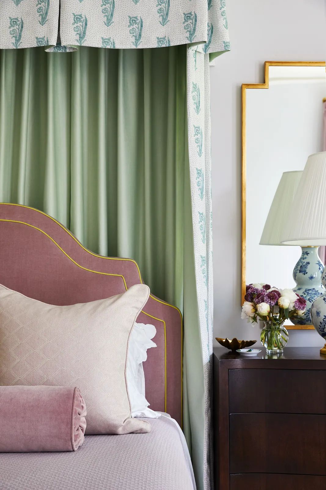 Bedroom with pink headboard, green drapery, side table with lamp, and vase of flowers.