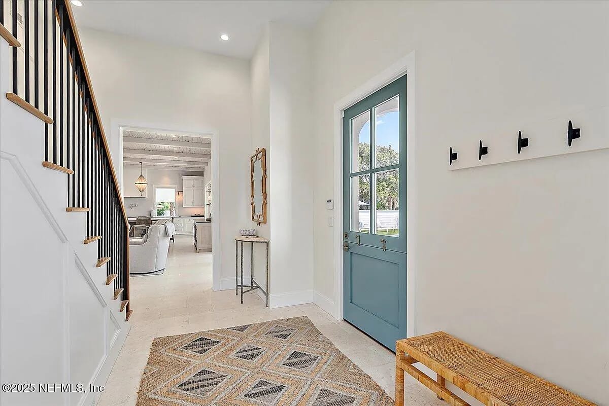 Foyer with blue door, woven bench, patterned rug, mirror, staircase, and wall hooks.