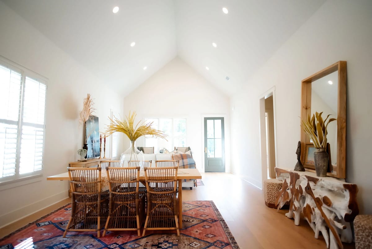 Dining room with wooden table, woven chairs, area rug, console table, plants, and large windows.