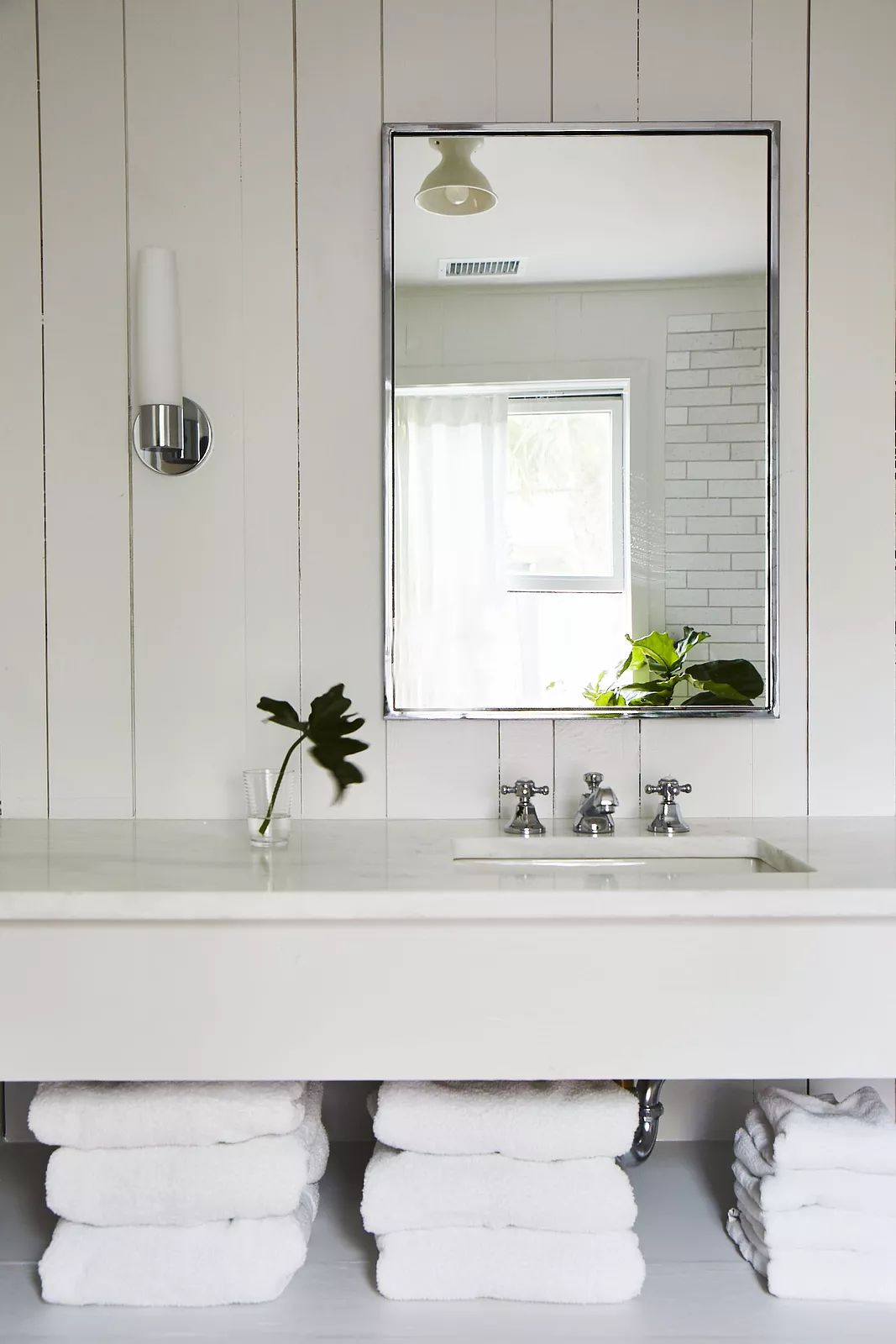 Bathroom with wall-mounted sink, chrome faucets, mirror, stacked towels, window, and plant.