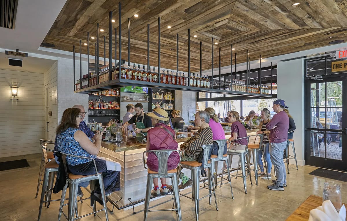 Dining room with bar, high stools, wood plank ceiling, concrete flooring, and patrons seated.