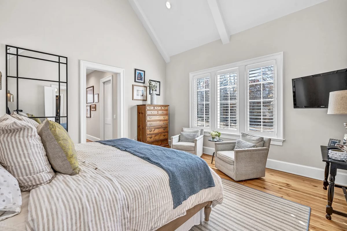 Bedroom with striped comforter, accent chairs, dresser, television, and vaulted ceilings.