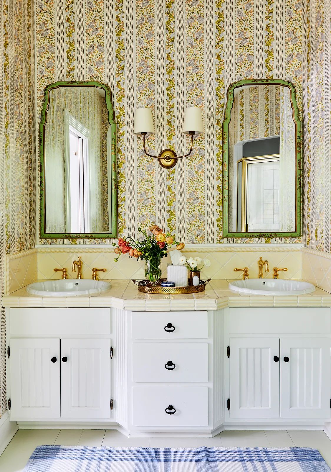 Bathroom with double sink vanity, floral wallpaper, mirrors, gold faucets, and a light blue rug