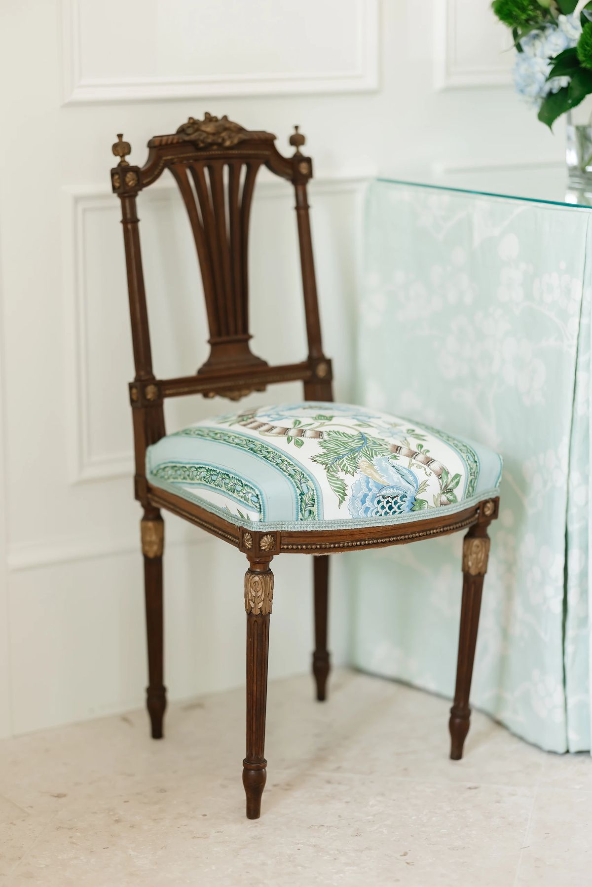 Wooden chair with floral cushioned seat near a glass table with light blue cover and white paneled walls.