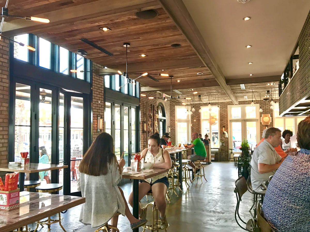 Dining area with high tables, metal stools, large windows, exposed brick walls, and wooden beams.