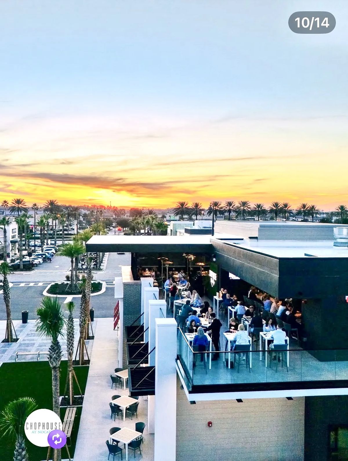 Outdoor dining area on a balcony with tables, palm trees, and a sunset view.