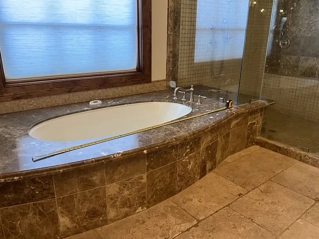 Bathroom with oval bathtub, granite countertop, glass shower, and beige stone tiles