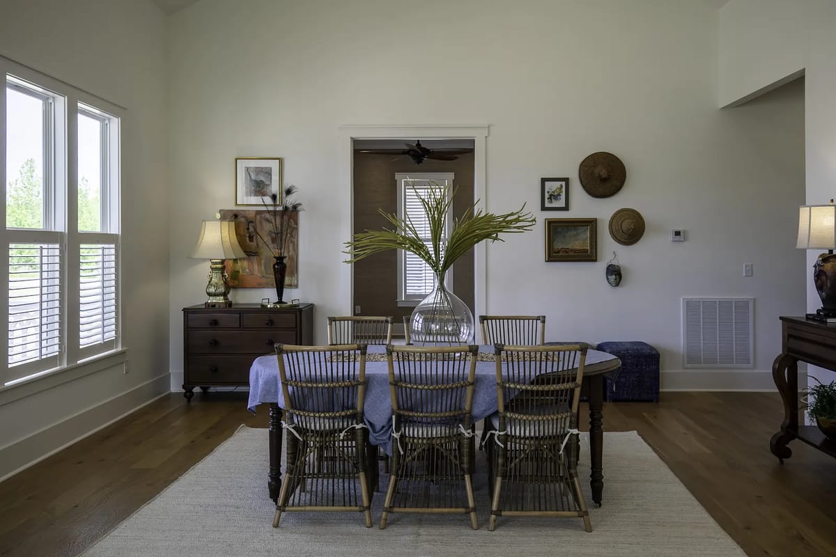 Dining room with wooden table, rattan chairs, vase of greenery, lamps, and framed artworks.