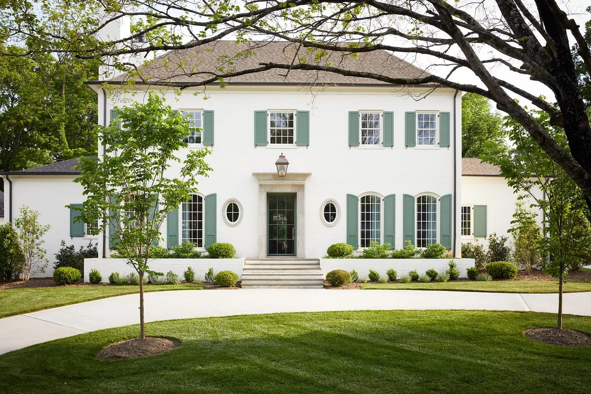 Front exterior of a white brick house with green shutters, windows, and manicured landscaping.
