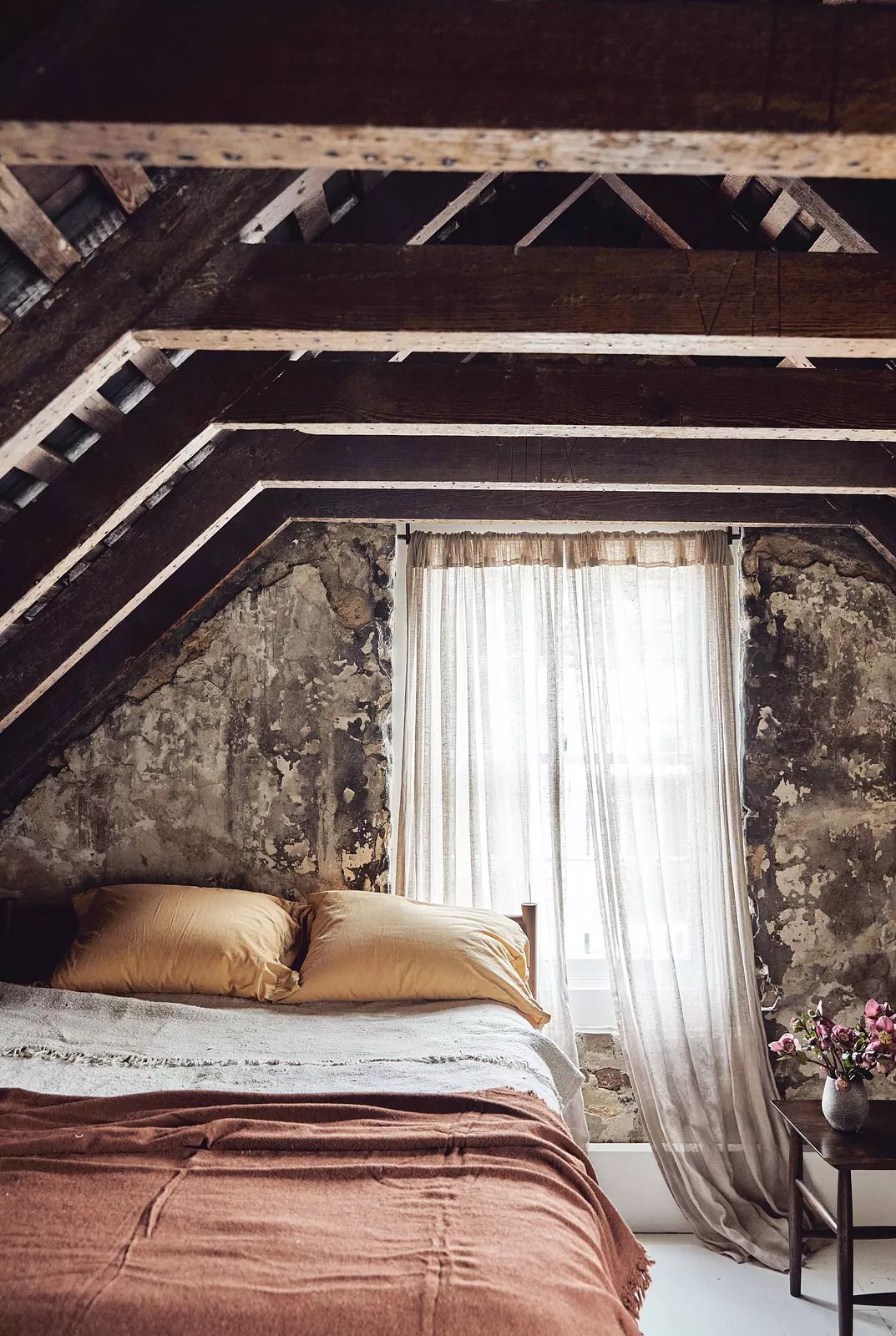Attic room with wooden beams, bed, sheer curtains, and small vase of flowers on bedside table.