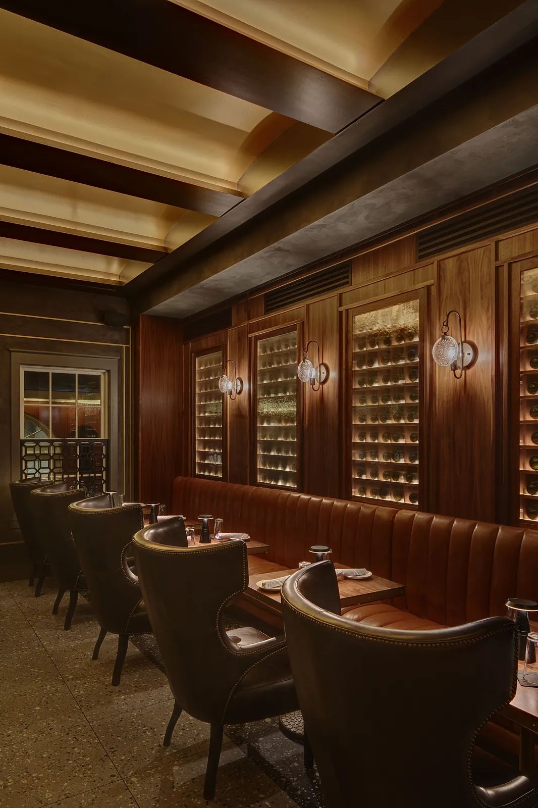 Dining room with leather banquette, wooden tables, dark wood paneling, wine shelves, and wall-mounted light fixtures.