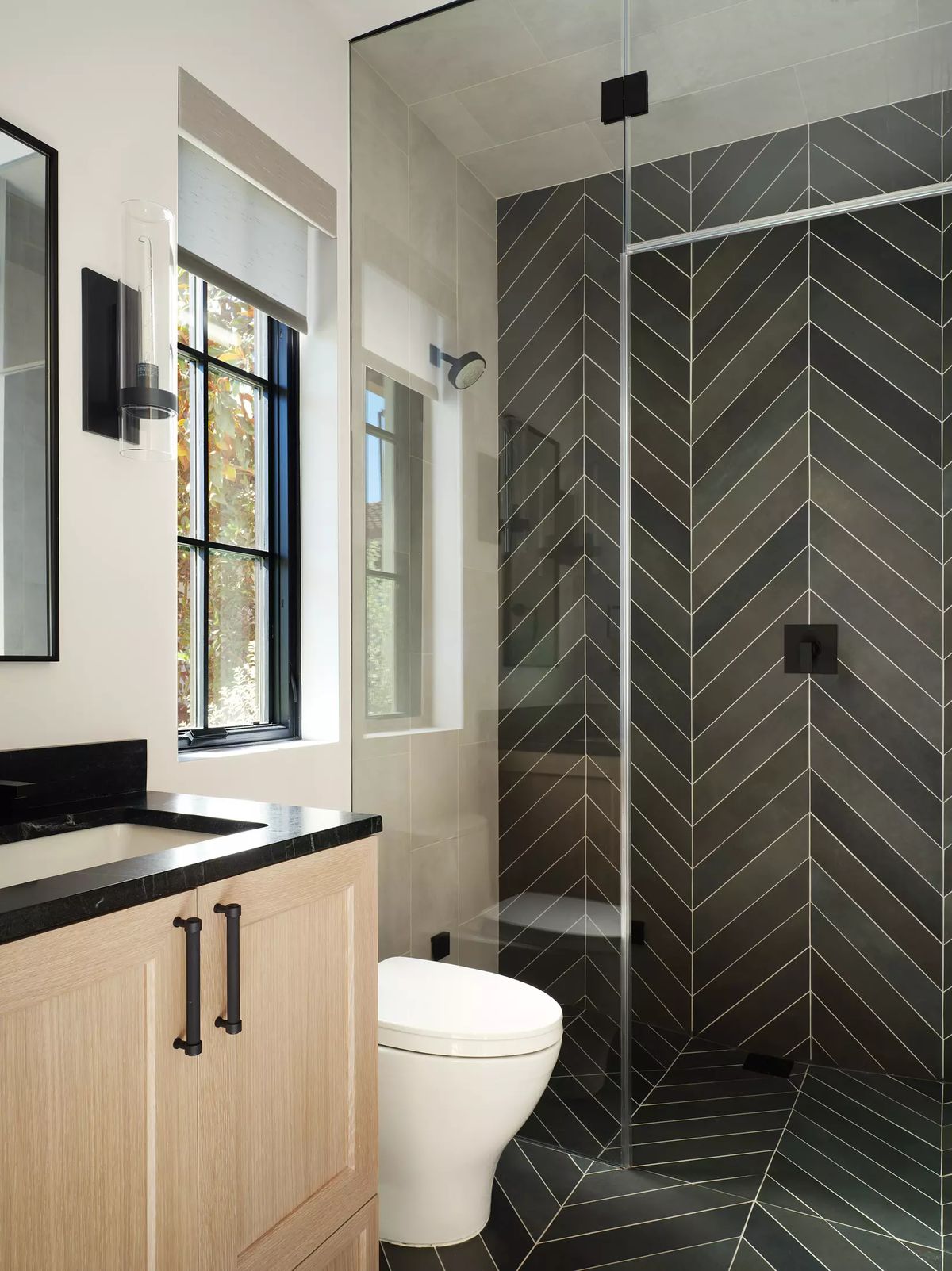 Bathroom with walk-in shower, wooden vanity, black countertop, dark tile, and window for natural light