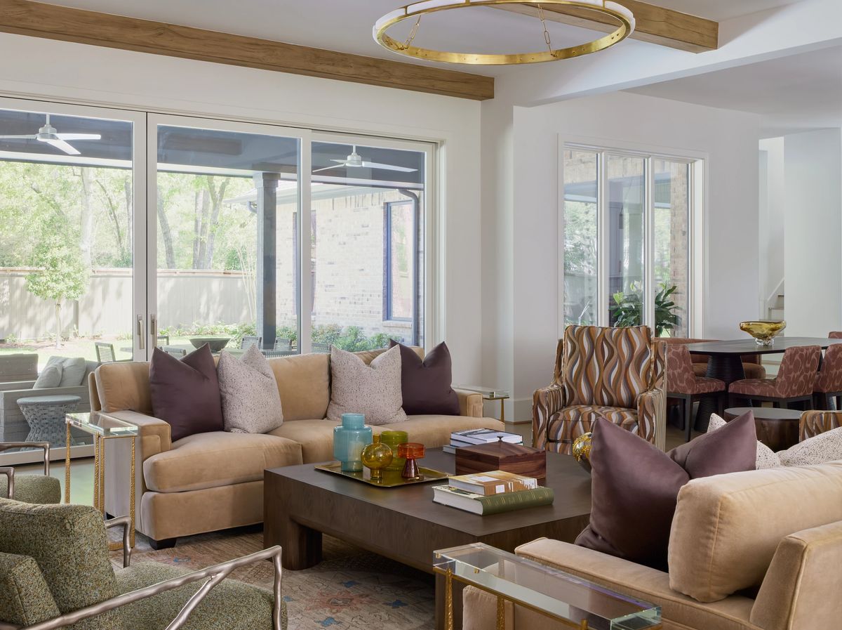 Living room with tan sectional sofa, brown coffee table, accent chairs, and large windows overlooking greenery.