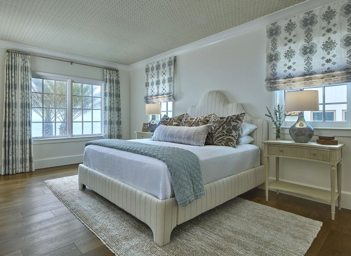 Bedroom with white bed, blue quilt, nightstands, lamps, and large windows with patterned curtains