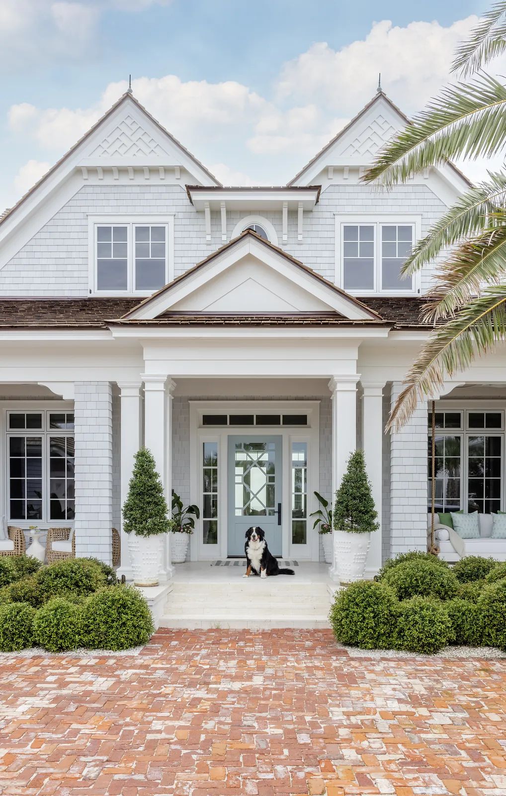 Front exterior of a house with double doors, potted plants, brick pathway, and porch with seating.