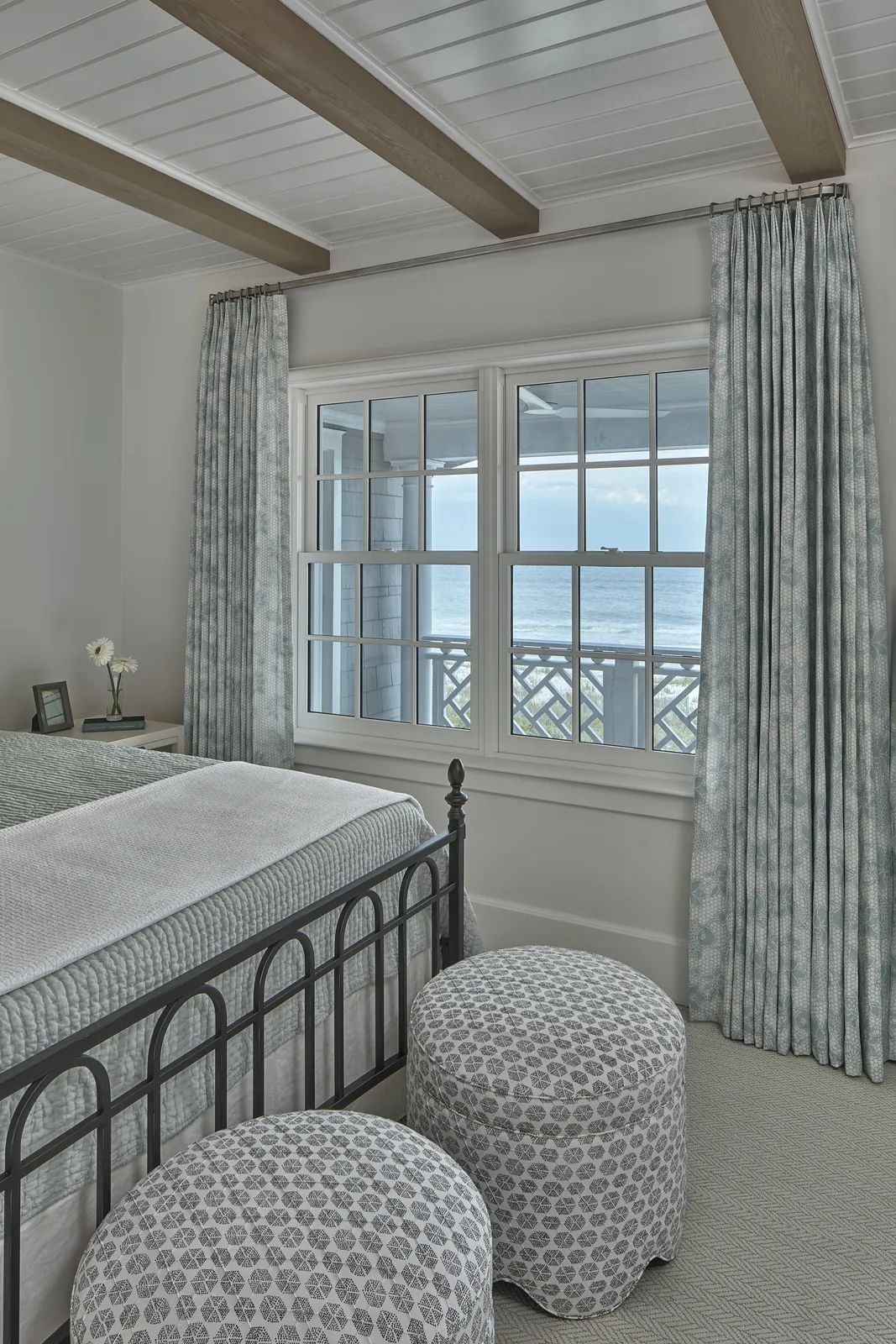 Bedroom with black metal bed frame, upholstered stools, sliding windows, and patterned drapes.