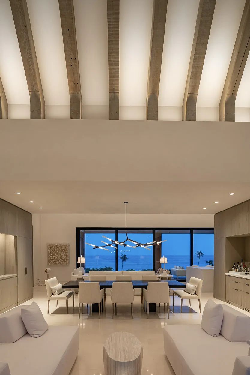 Living room with white sofas, circular table, dining table, white chairs, large window, and wooden beams ceiling.